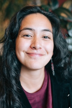 A smiling portrait of an Indigenous Alaskan woman indoors, plants visible in the background. Shot in Anchorage, Alaska, USA.